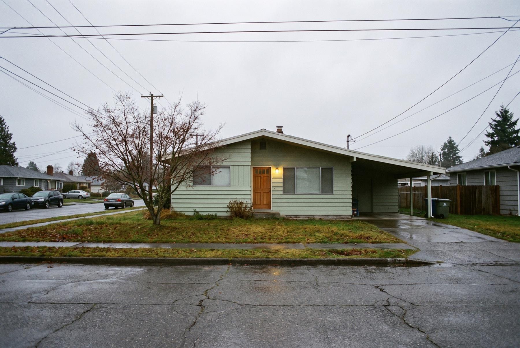 A mid-century rambler with a carport on a flat street on a gray winter morning, porch light on