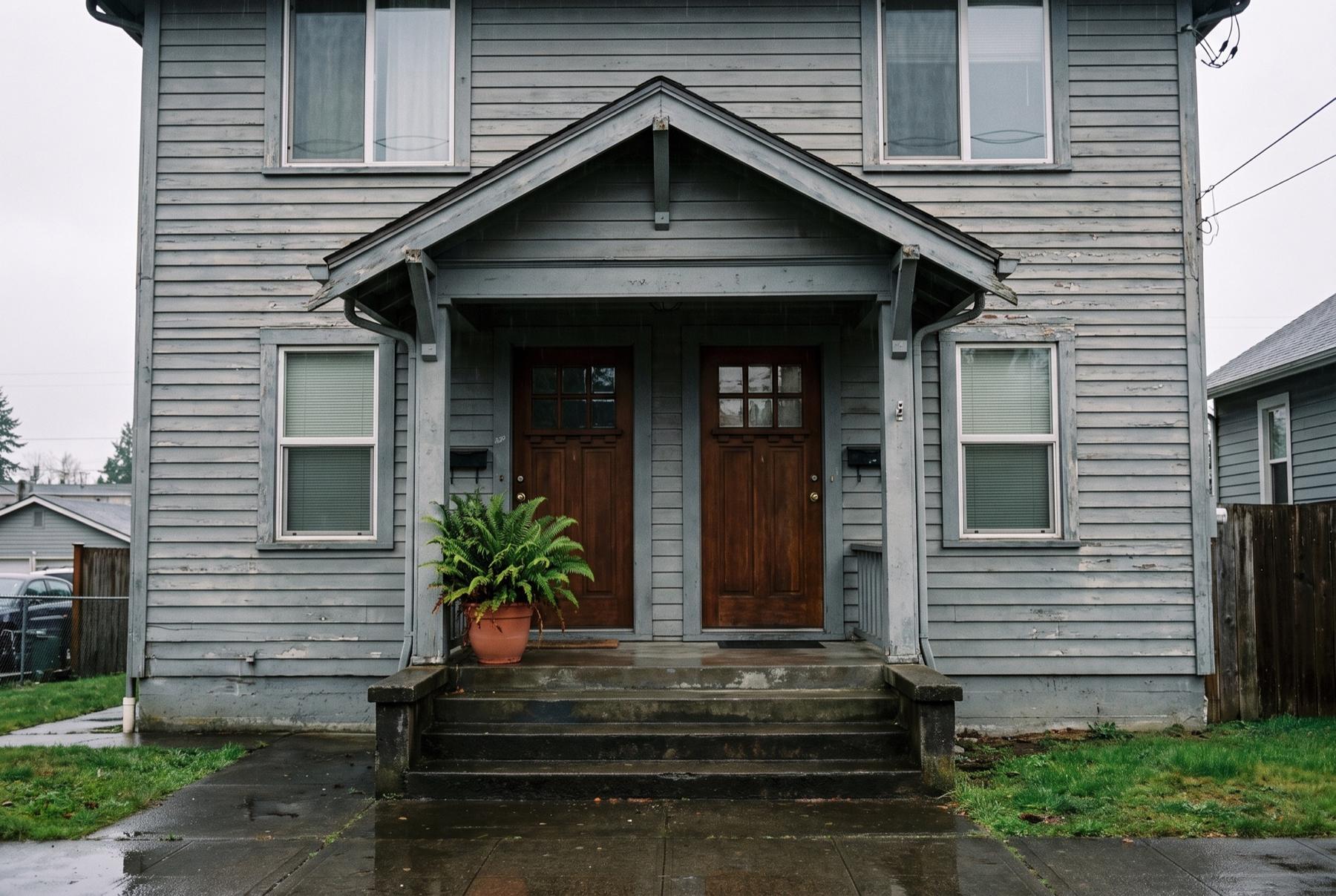 An older gray duplex with two wooden front doors under one porch roof, the steps wet from rain