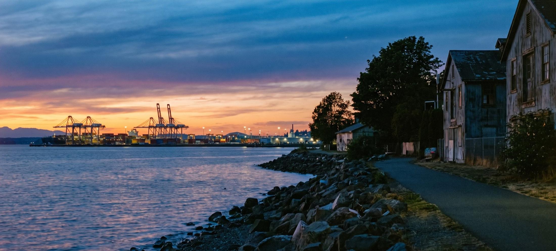 Paved waterfront path beside a rock bulkhead and weathered wooden buildings at dusk, with lit container cranes across the bay